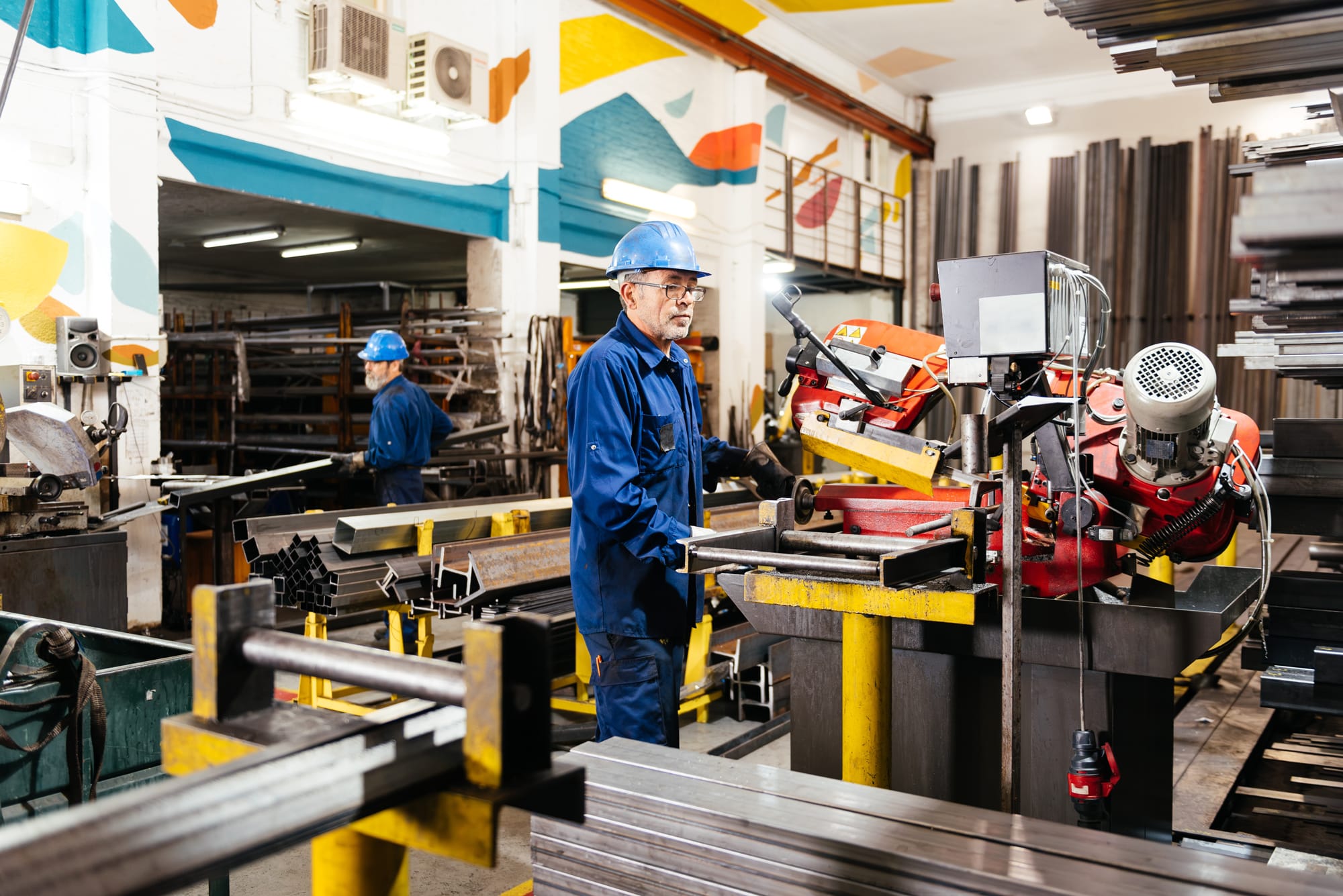 Two manufacturing workers in blue hard hats and uniforms operating bandsaw in vibrant industrial workshop, showcasing teamwork for temp-to-perm success.