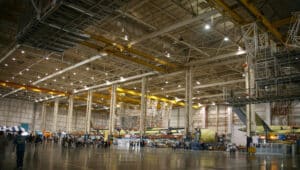Spacious aerospace manufacturing plant with yellow overhead cranes, aircraft fuselage parts, steel beams, and workers in hangar.