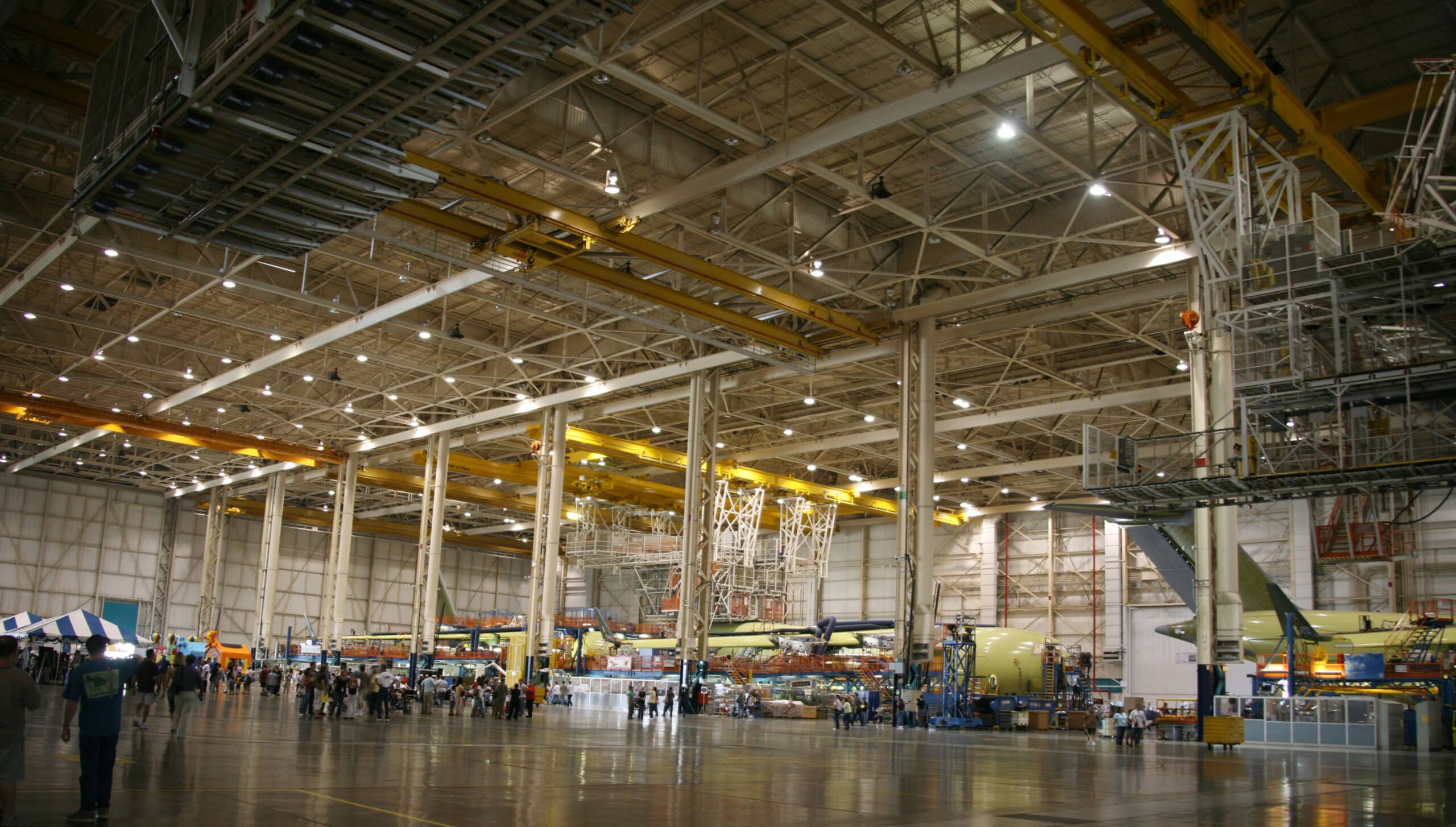 Spacious aerospace manufacturing plant with yellow overhead cranes, aircraft fuselage parts, steel beams, and workers in hangar.