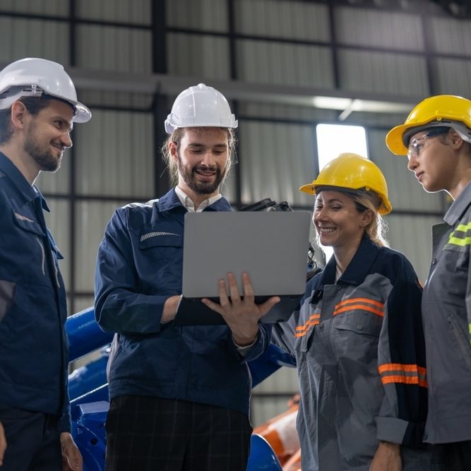 A diverse group of smiling industrial professionals collaborates over a laptop on the factory floor