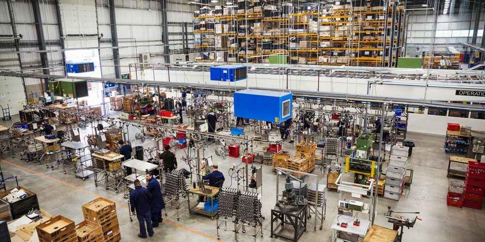 Busy manufacturing assembly floor with organized workstations, workers in uniforms, tall storage shelves, and overhead equipment.