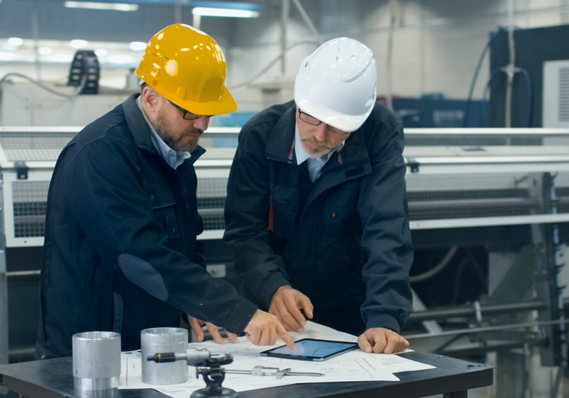 Two engineers discuss a blueprint while checking information on a tablet computer in a factory Two engineers discuss a blueprint while checking information on a tablet computer in a factory