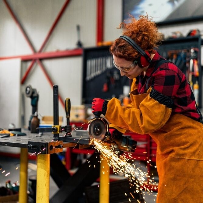 Woman in STEM using grinder to cut a metal bar, wearing gloves and a apron with protective sleeves Woman in STEM using grinder to cut a metal bar, wearing gloves and a apron with protective sleeves