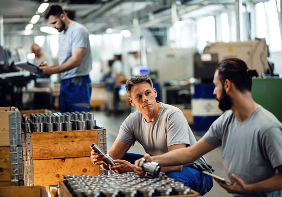 Manufacturing team discussing metal parts over a wooden crate, with colleague taking notes on clipboard in busy factory.