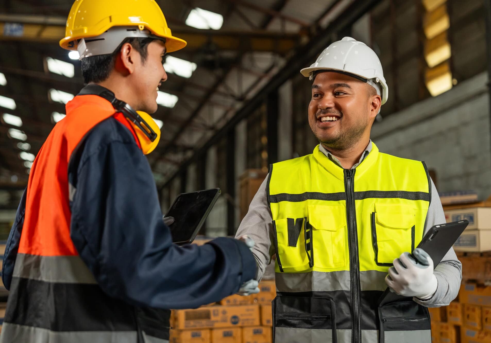 Two manufacturing workers in safety gear shaking hands with tablets in warehouse, symbolizing temp-to-perm teamwork and success.