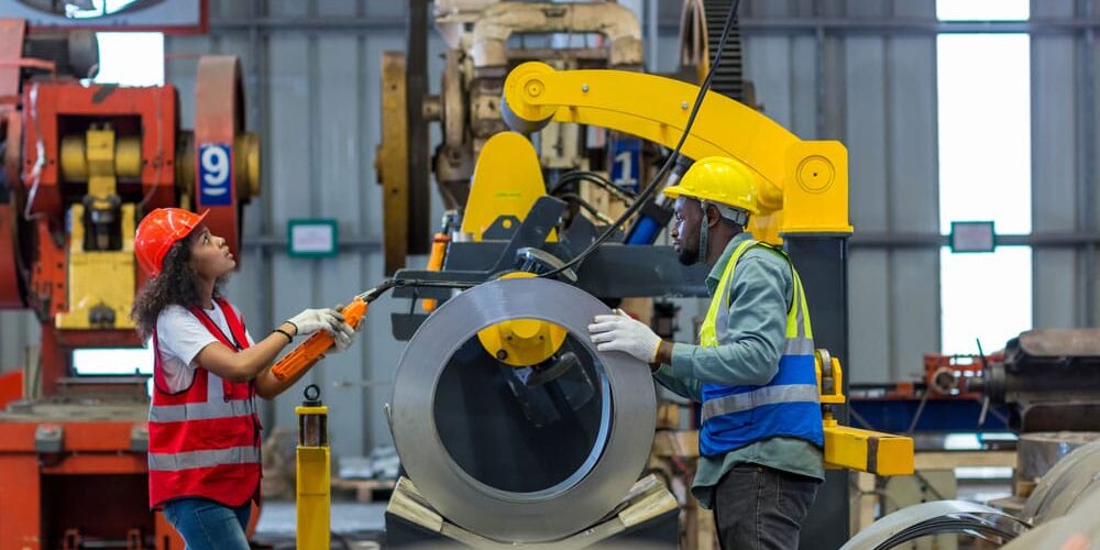 Female and male workers operating machinery to manage large metal coil in well-lit industrial manufacturing plant.