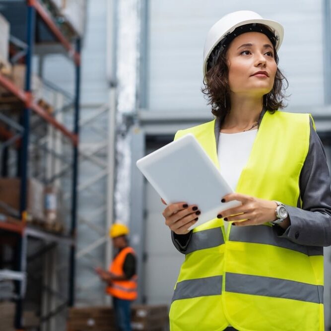 female warehouse supervisor in hard hat and safety vest holding digital tablet during work
