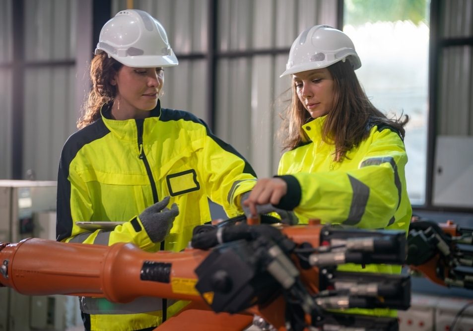 Engineers,Dressed,In,Safety,Gear,Are,Working,Together,On,A Engineers dressed in safety gear are working together on a robotic arm in a manufacturing plant.