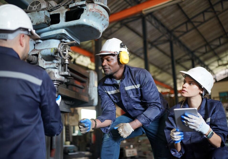 Three factory workers in white hard hats and blue uniforms inspecting and monitoring machinery with tablet in large industrial space.