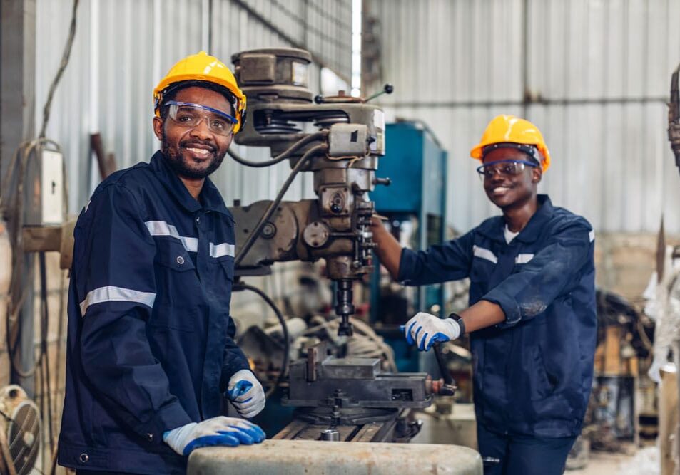 Two smiling workers in yellow hard hats, goggles, and blue uniforms operating drill press machinery in manufacturing workshop.